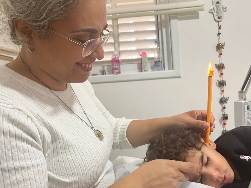 רשין מחזיקה נר הופי דולק באוזנו של ילד השוכב על צידו בקליניקה